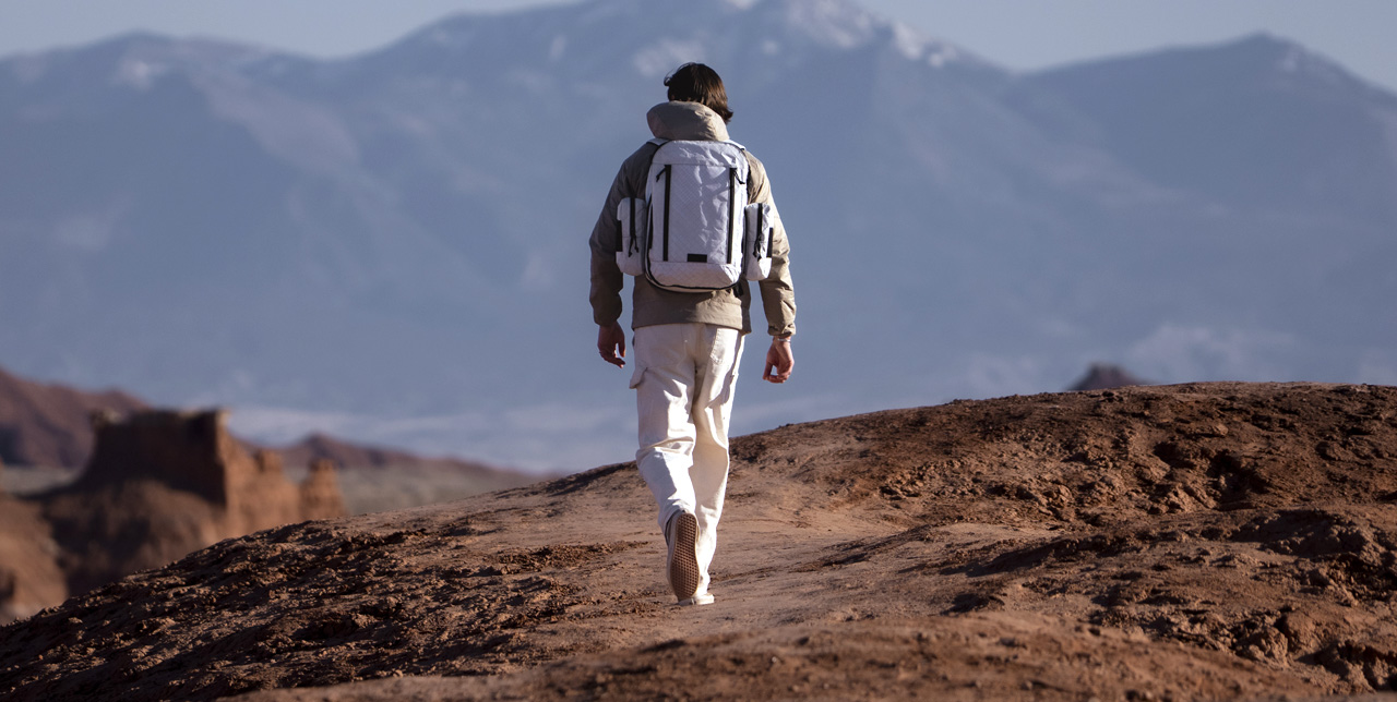 MAN WALKING IN DESERT WITH WHITE BACKPACK