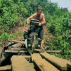 Explorer Jude Kriwald pushing his bike over a wooden bridge in Liberia