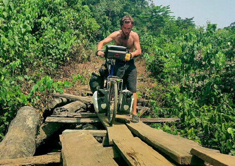 Explorer Jude Kriwald pushing his bike over a wooden bridge in Liberia