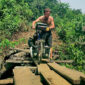 Explorer Jude Kriwald pushing his bike over a wooden bridge in Liberia