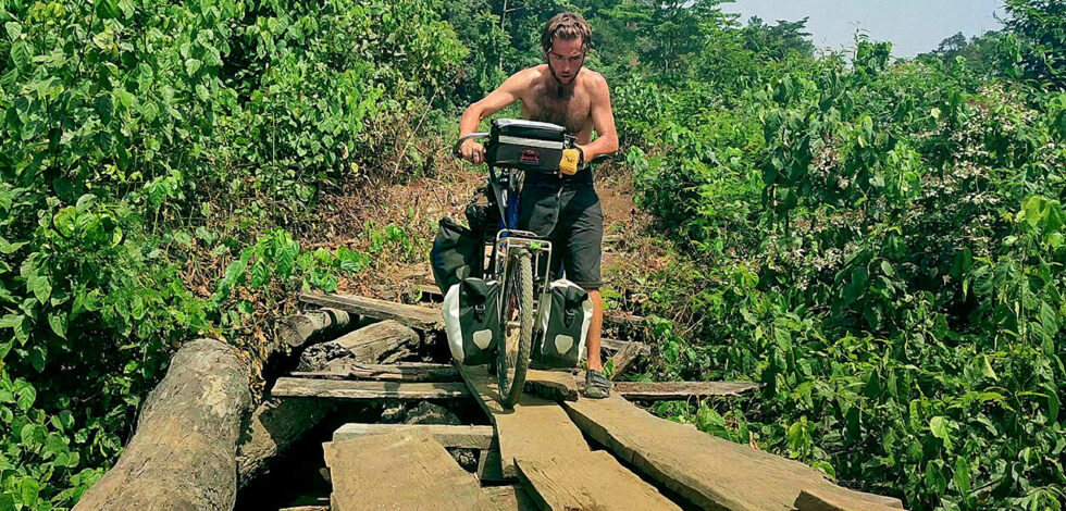 Explorer Jude Kriwald pushing his bike over a wooden bridge in Liberia