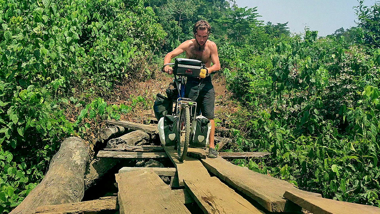 Explorer Jude Kriwald pushing his bike over a wooden bridge in Liberia